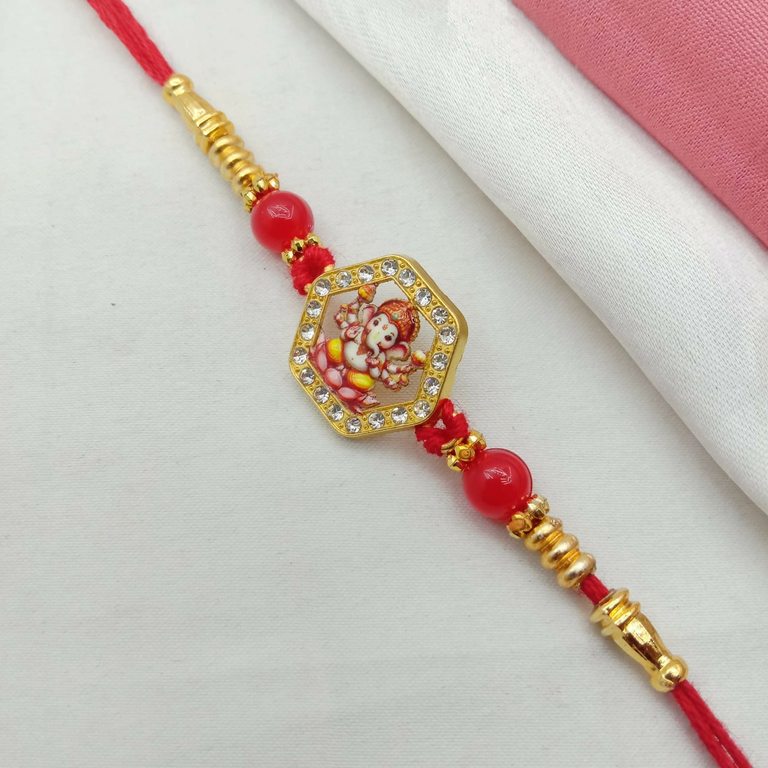 A red thread bracelet featuring a Ganesha figure charm with a rhinestone hexagon border and red beads is on a white cloth.