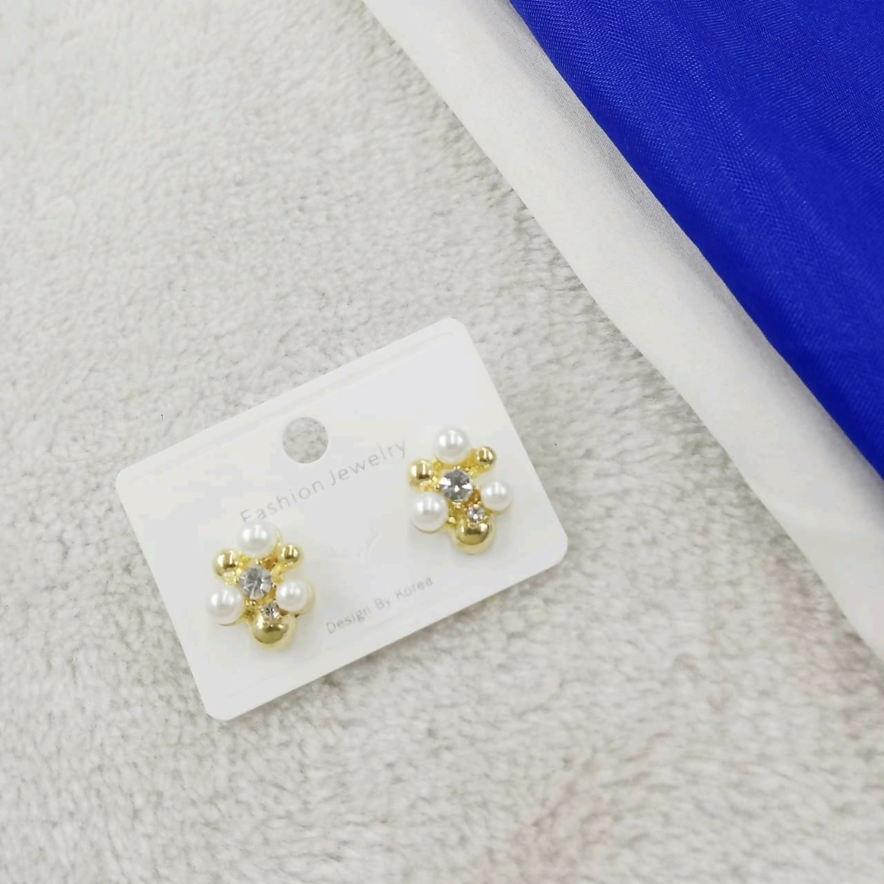 A pair of gold-plated floral stud earrings with crystals and mini pearls on a white card, placed on a textured white surface