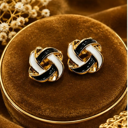 A pair of black and white enamel knot stud earrings with a gold-tone base, displayed on a velvet surface.