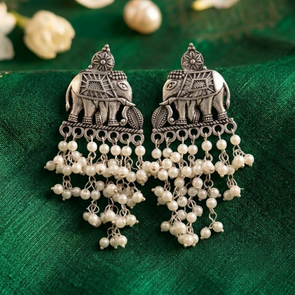 A pair of oxidized silver elephant earrings with white pearl drops on a green fabric background.