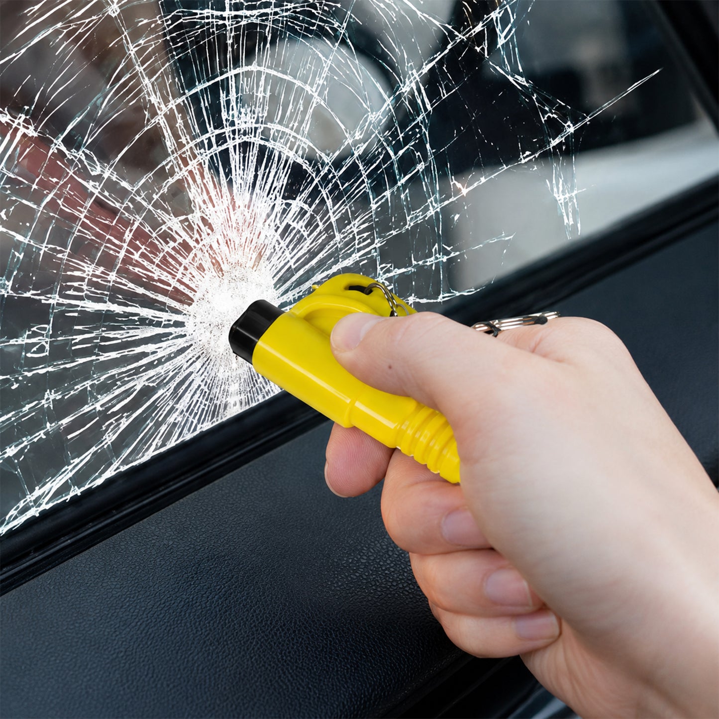 A hand firmly presses a yellow car safety hammer against a car window, creating a spiderweb crack pattern in the glass.