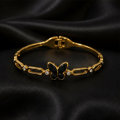 The gold-tone bracelet with a black butterfly charm and crystal accents is displayed on a black satin surface.
