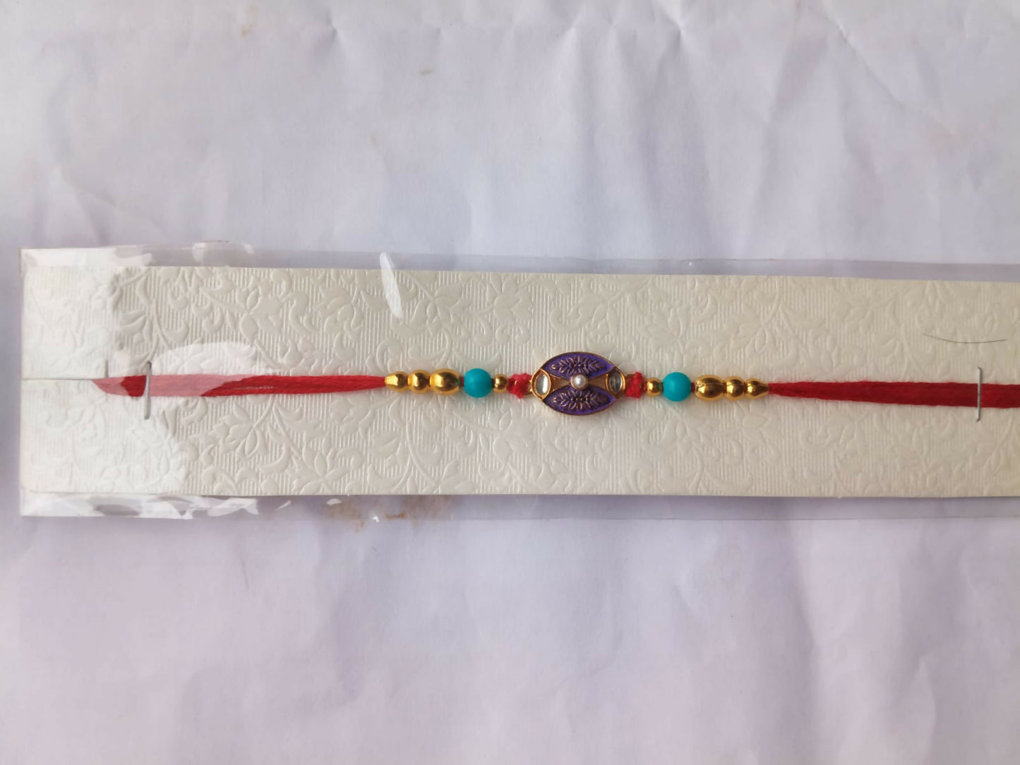 The red thread with a purple and gold oval charm, gold, and turquoise beads is displayed in clear packaging on a white