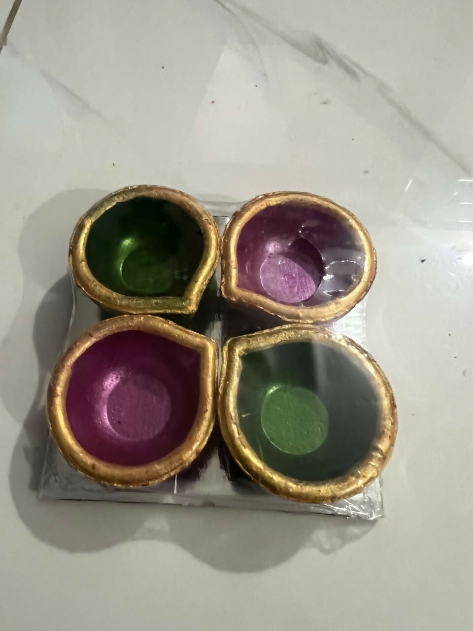 Four clay diyas, two green and two magenta, with golden rims, packaged in plastic on a tiled surface.