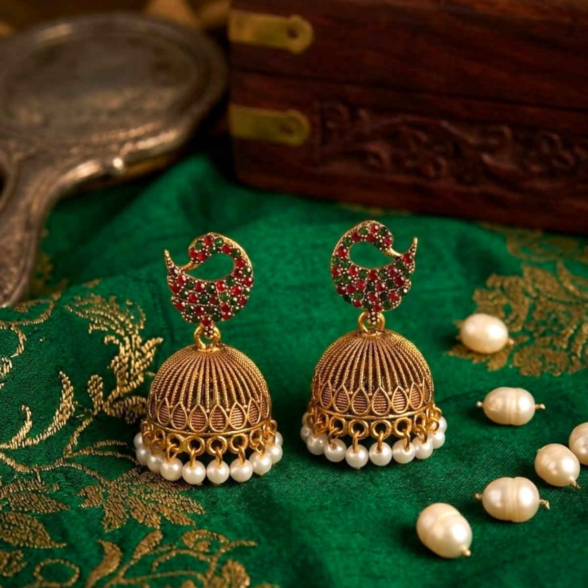 A pair of gold jhumka earrings with red and green stones and pearl drops on a green fabric.