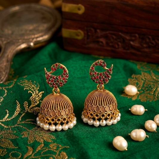 A pair of gold jhumka earrings with red and green stones and pearl drops on a green fabric.
