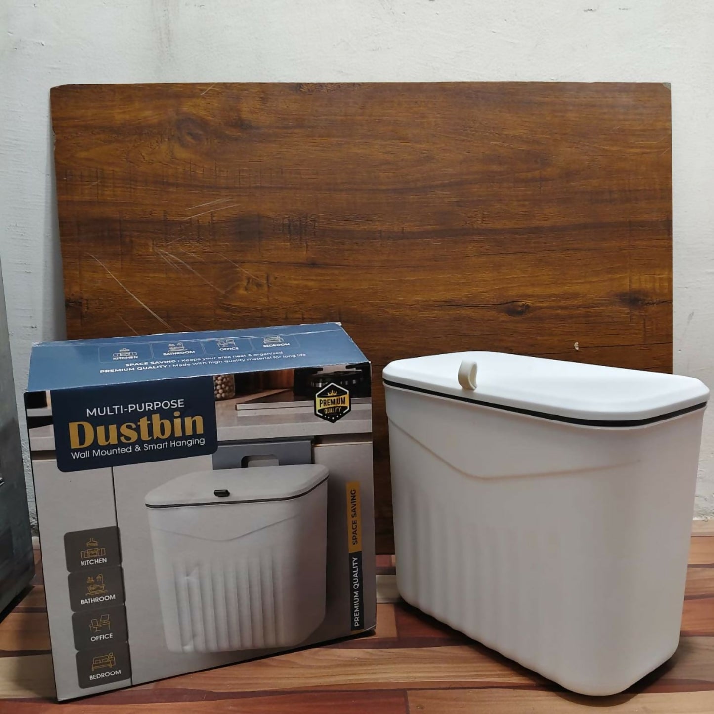 A white plastic dustbin and its blue and white product box are displayed on a light brown floor next to a wooden board.