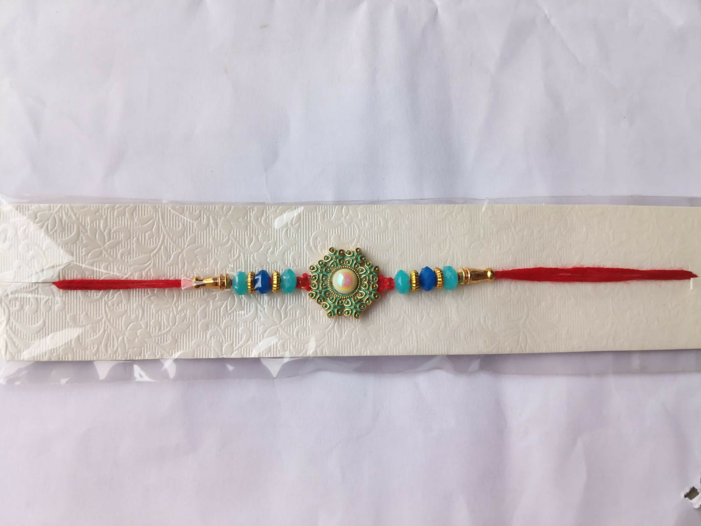 A red thread with a teal and gold medallion, featuring a central pearl, and blue beads is packaged on a white card.