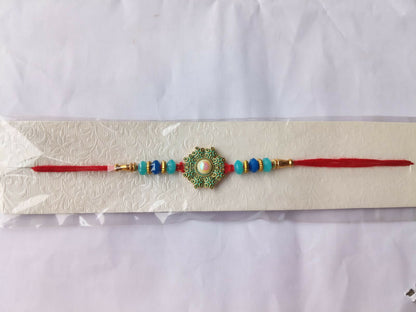 A red thread with a teal and gold medallion, featuring a central pearl, and blue beads is packaged on a white card.