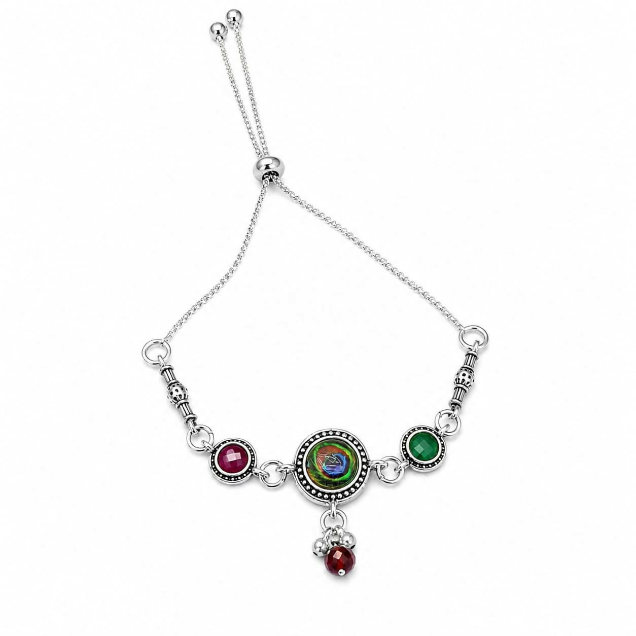 An antique silver adjustable bracelet with a peacock eye cabochon and maroon and green side stones is centered on a white
