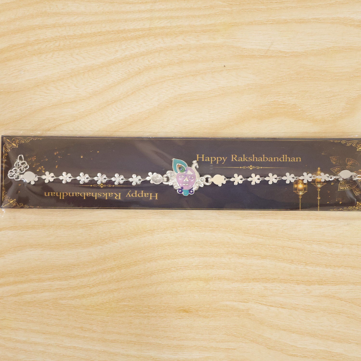 The silver rakhi with Krishna face charm displayed in its clear plastic packaging on a wooden surface.