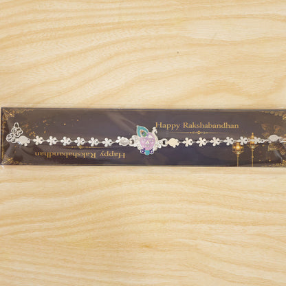 The silver rakhi with Krishna face charm displayed in its clear plastic packaging on a wooden surface.