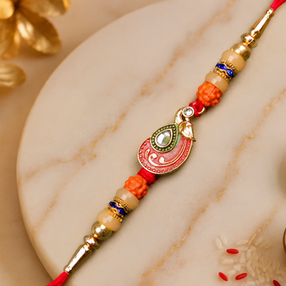 A red and gold bracelet with a peacock charm, orange and cream beads, and blue accents, rests on a marble surface with