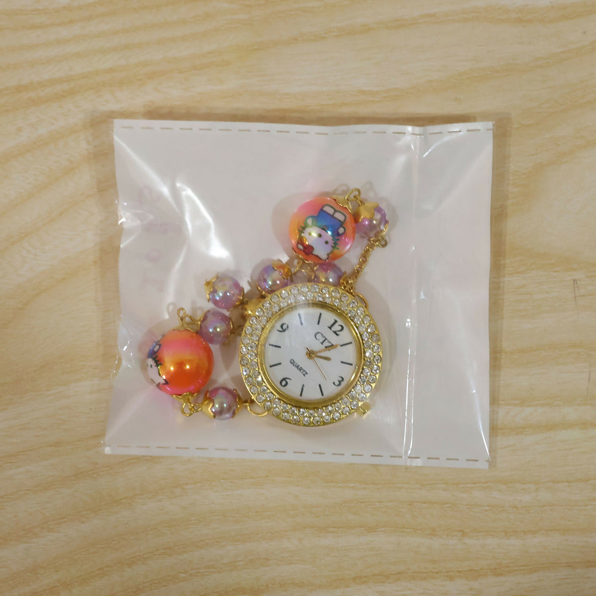 A gold-tone bracelet watch with a rhinestone dial, orange cartoon character beads, and smaller purple beads, sealed inside a