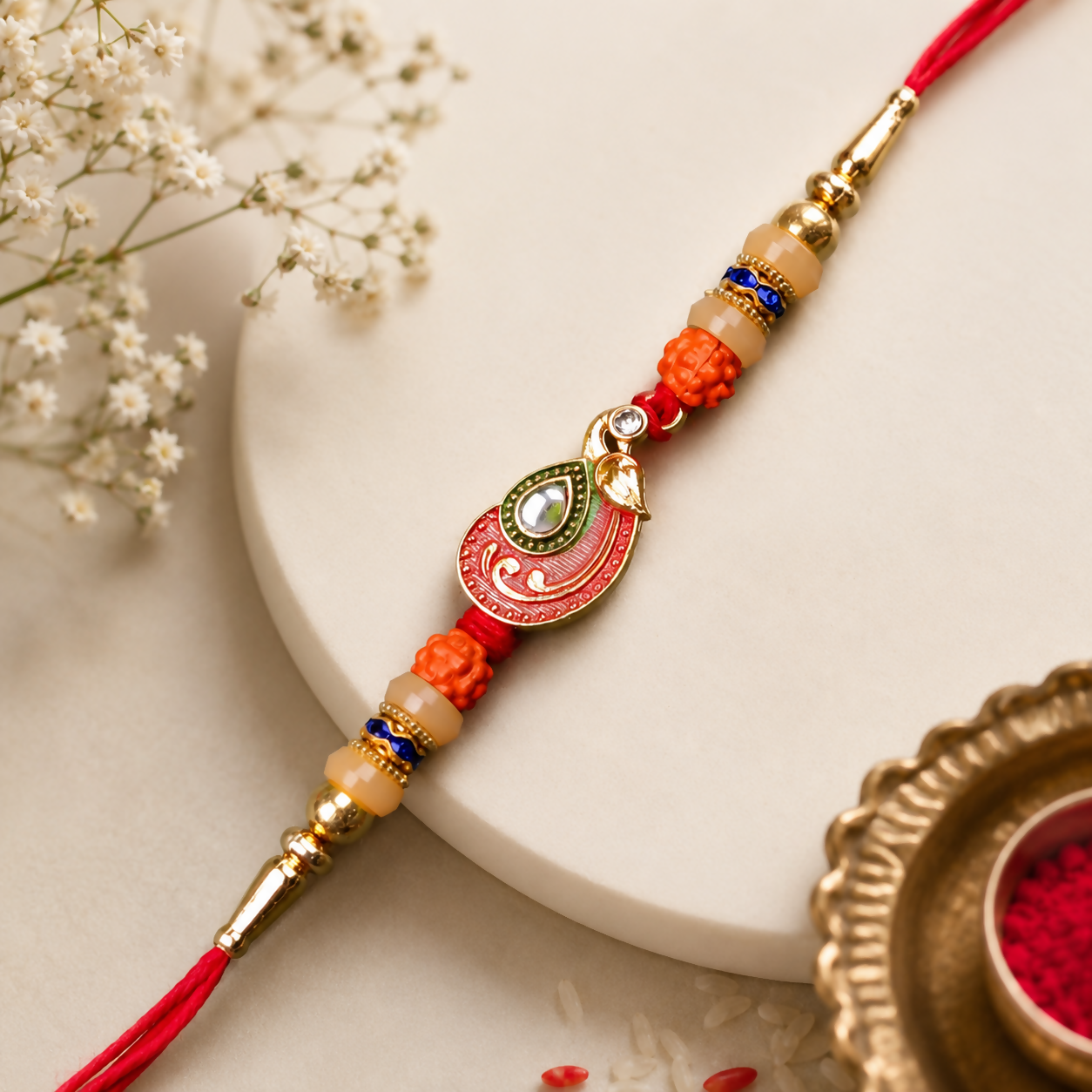 A red and gold bracelet with a peacock charm, orange and cream beads, and blue accents, rests on a light cream surface with