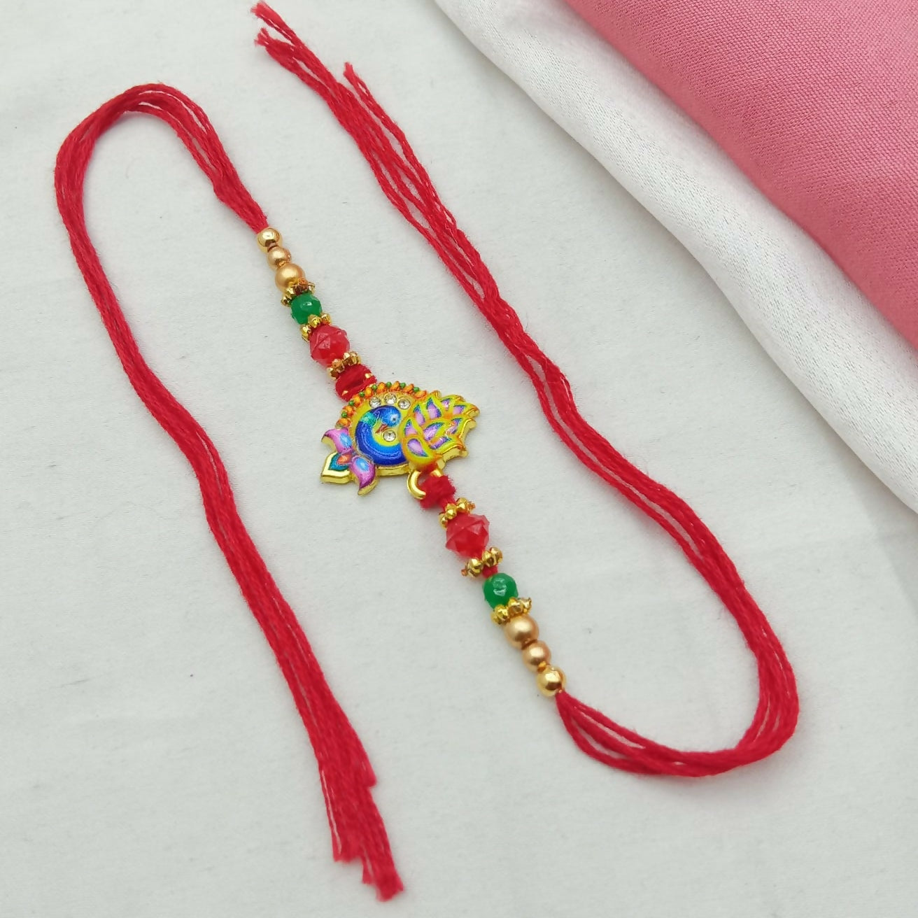 A golden-plated peacock enamel design with crystal stones and beaded thread is laid flat on a white and pink fabric