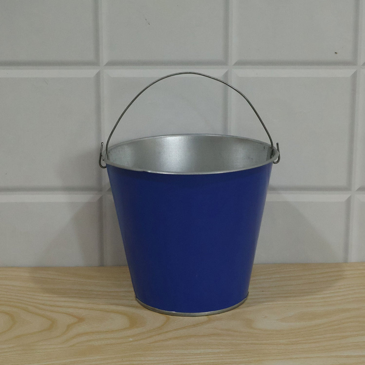 A blue metal bucket with a silver handle and rim, resting on a light wooden surface against a white tiled wall.