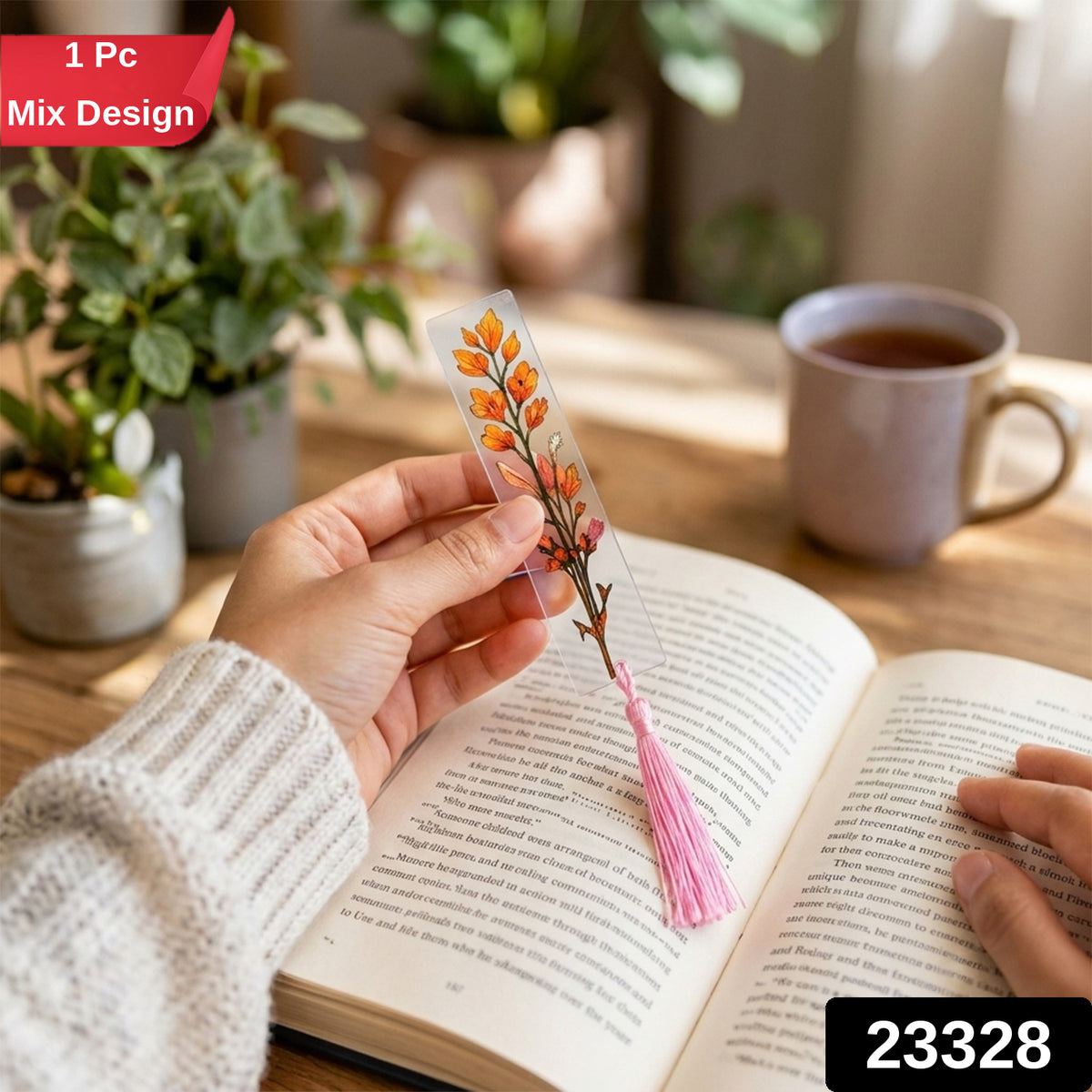 A hand holds a transparent bookmark with an orange leafy design and a pink tassel, with plants and a cup.
