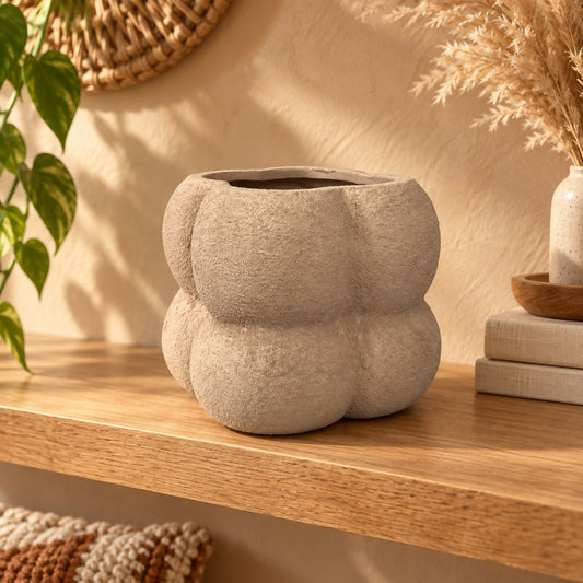Textured beige ceramic planter with a unique rounded shape sits on a wooden shelf next to a green plant.