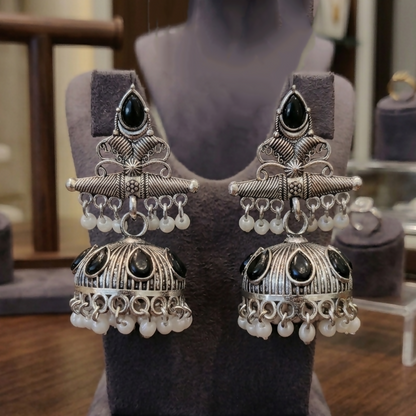 A pair of oxidized silver jhumka earrings with black stones and white pearls is displayed on  grey bust-style jewelry