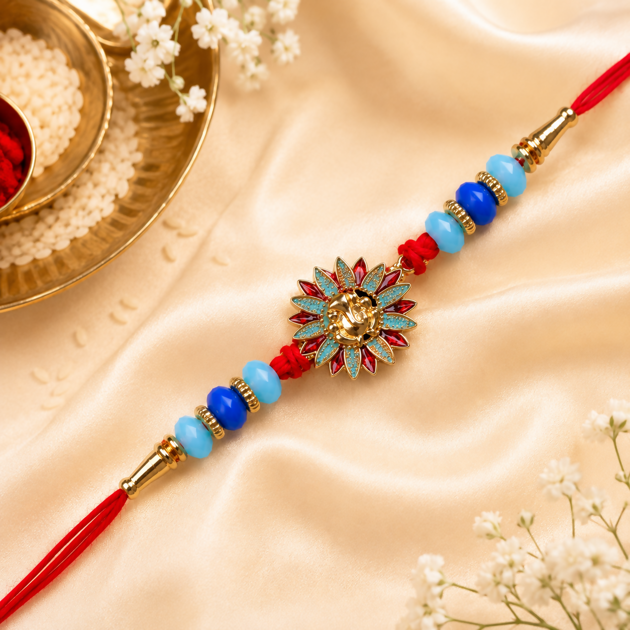 A red thread bracelet with a gold Ganesh charm, blue and light blue beads, and gold accents, on a cream satin fabric with