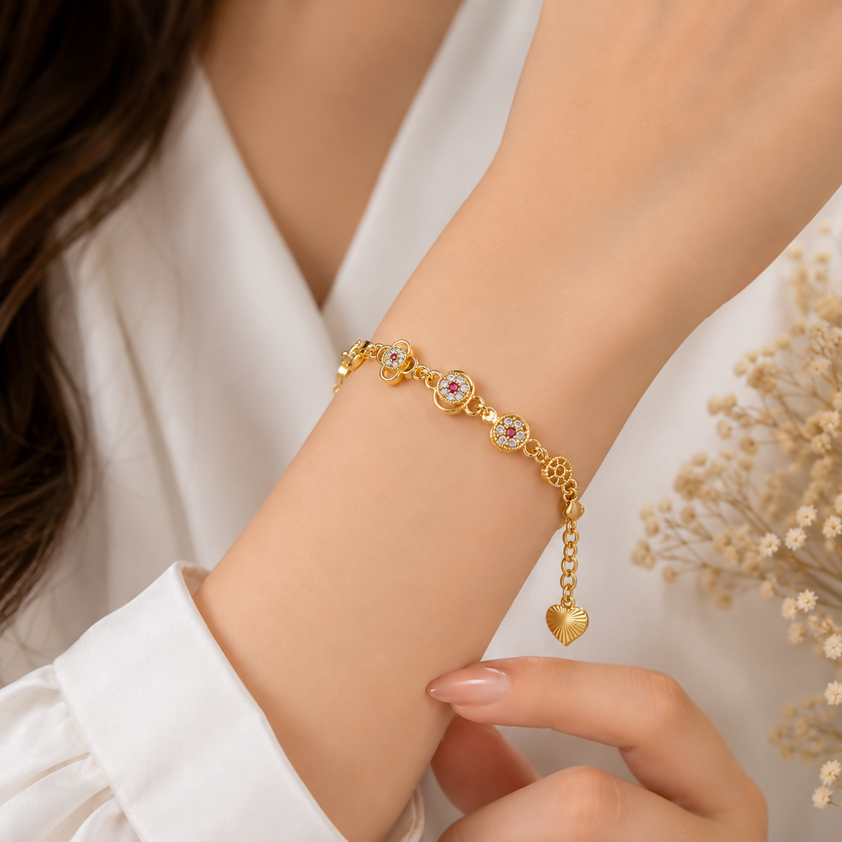 A woman's wrist wears a gold-plated bracelet with floral charms, red, and clear gems, against a white shirt.
