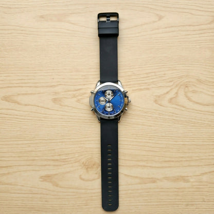 Top-down view of a black silicone strap watch with a silver case and blue dial, on a light wooden surface.