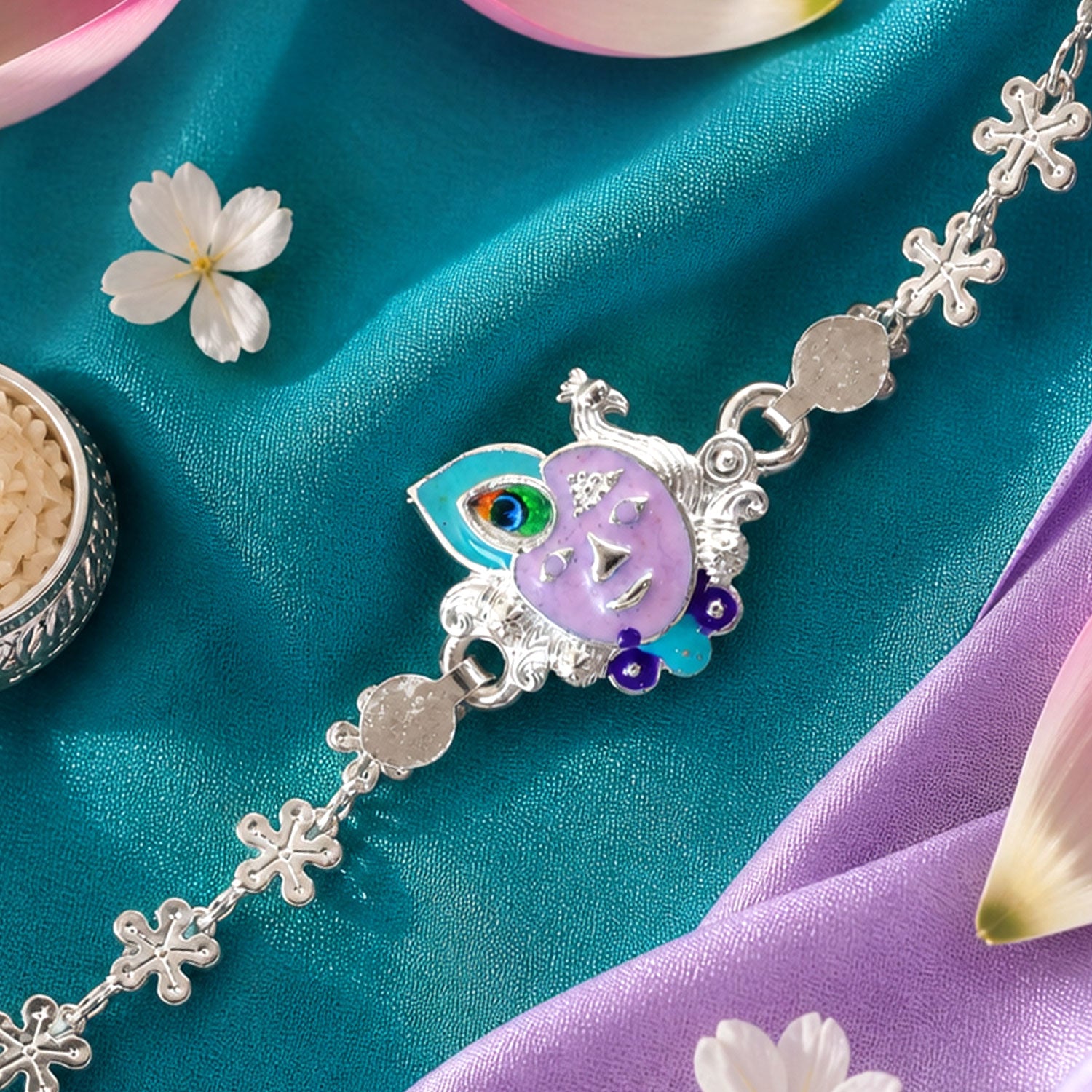 Close-up of a silver rakhi with a purple enamel Krishna face and peacock feather charm on teal and purple fabric.