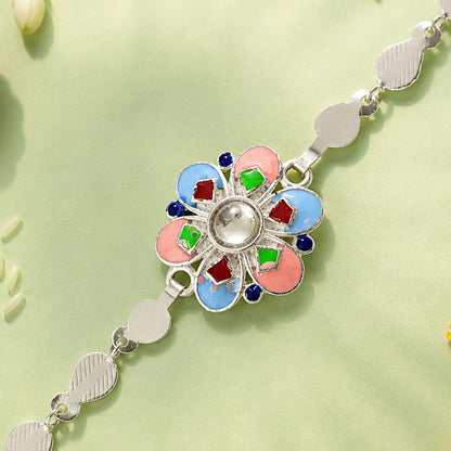 A silver chain rakhi with a colorful enamel flower charm on a light green background with rice grains.