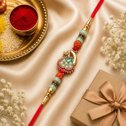 A gold peacock-shaped Rakhi with green and pearl details, flanked by orange and mint green beads, on a red thread.