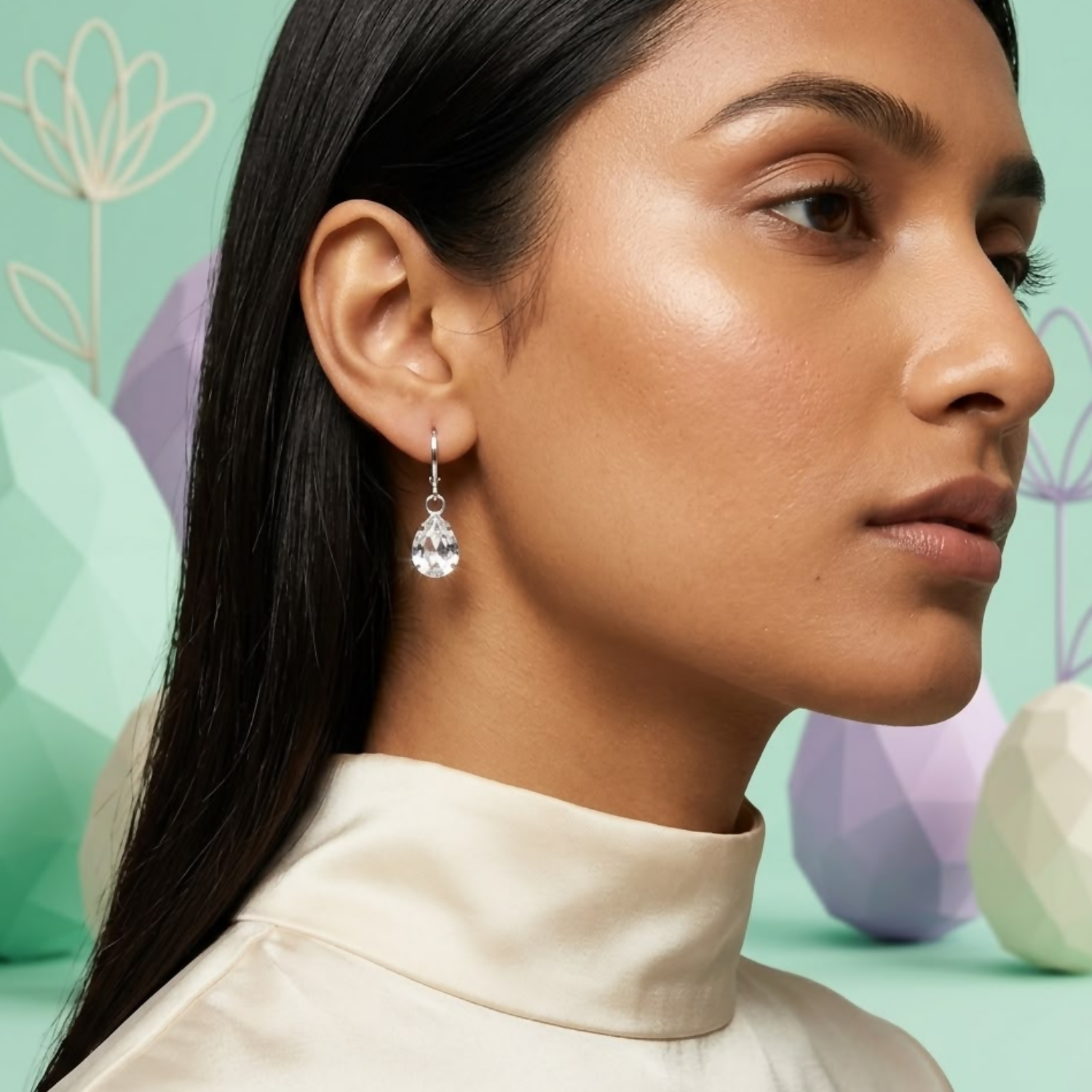 A woman wears a silver teardrop crystal earring, with pastel geometric shapes in the background.