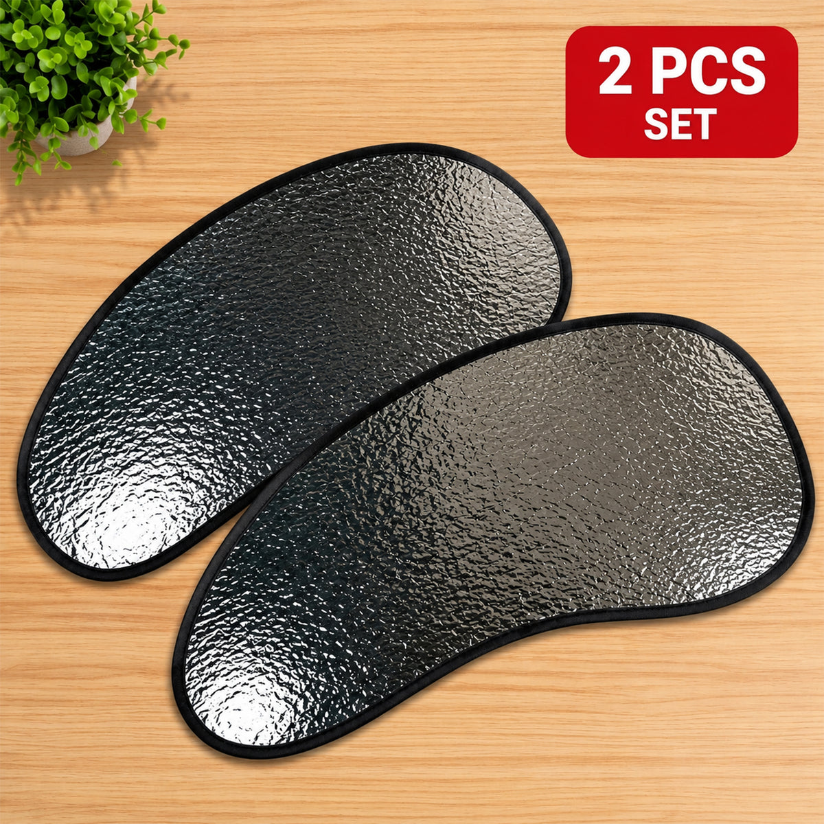 Two silver reflective car sunshades with black edges lying on a wooden surface next to a small plant.