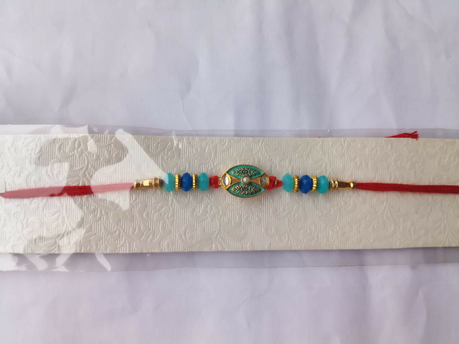 A red thread bracelet with blue and light blue beads and a central golden oval charm with a light blue design, is displayed