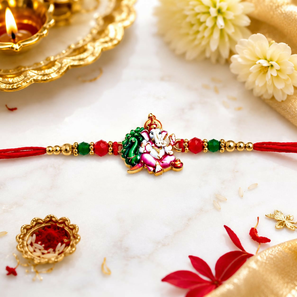 A red thread with an enamel Ganesha charm, green and red crystal beads, and golden beads, on a marble surface with flowers