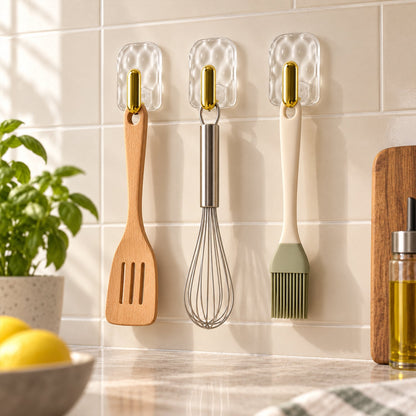 Three clear plastic hooks with gold accents hold a wooden spatula, a metal whisk, and a silicone brush on a white tiled wall.