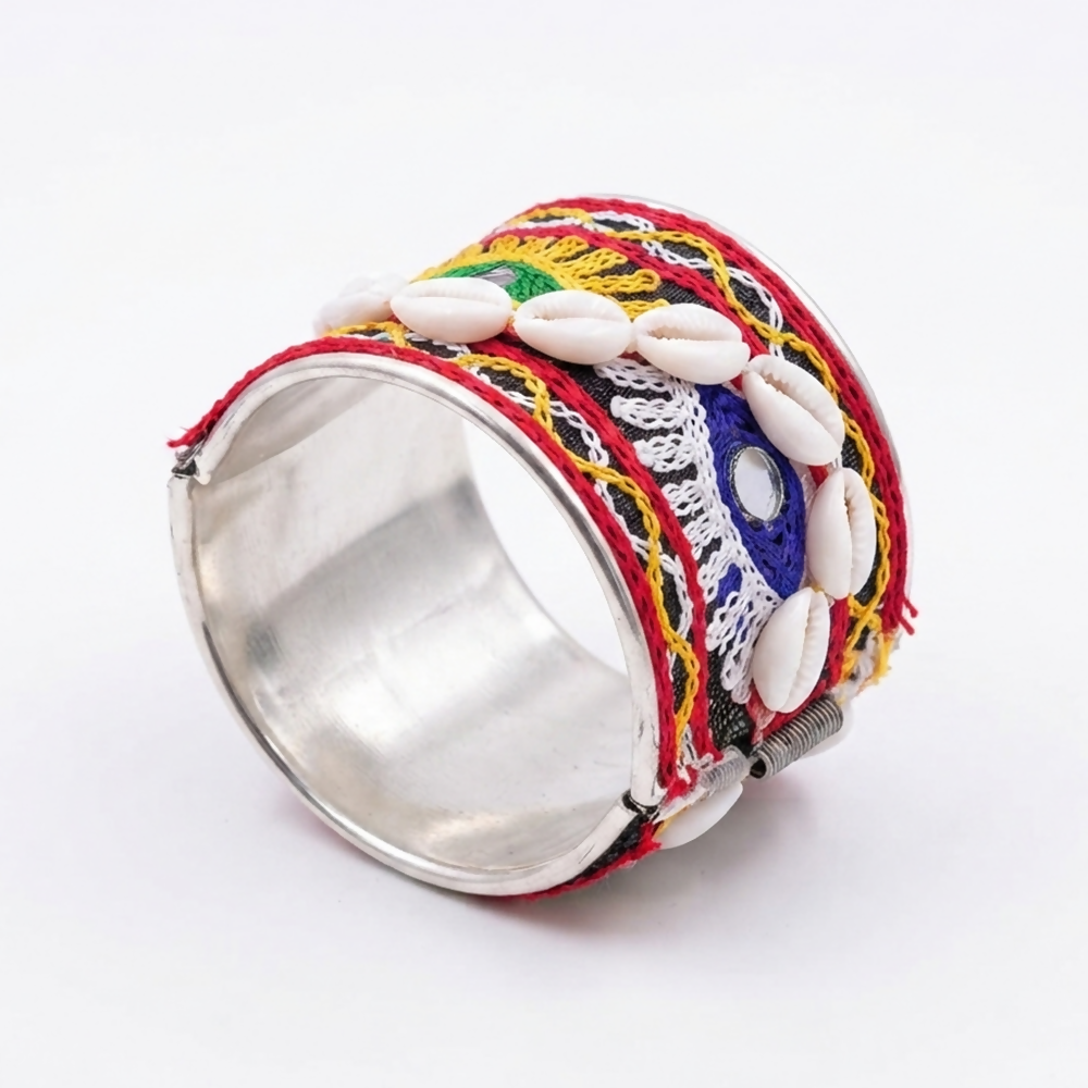 A wide silver bangle with black fabric, red, yellow, and blue embroidery, and cowrie shells on white.