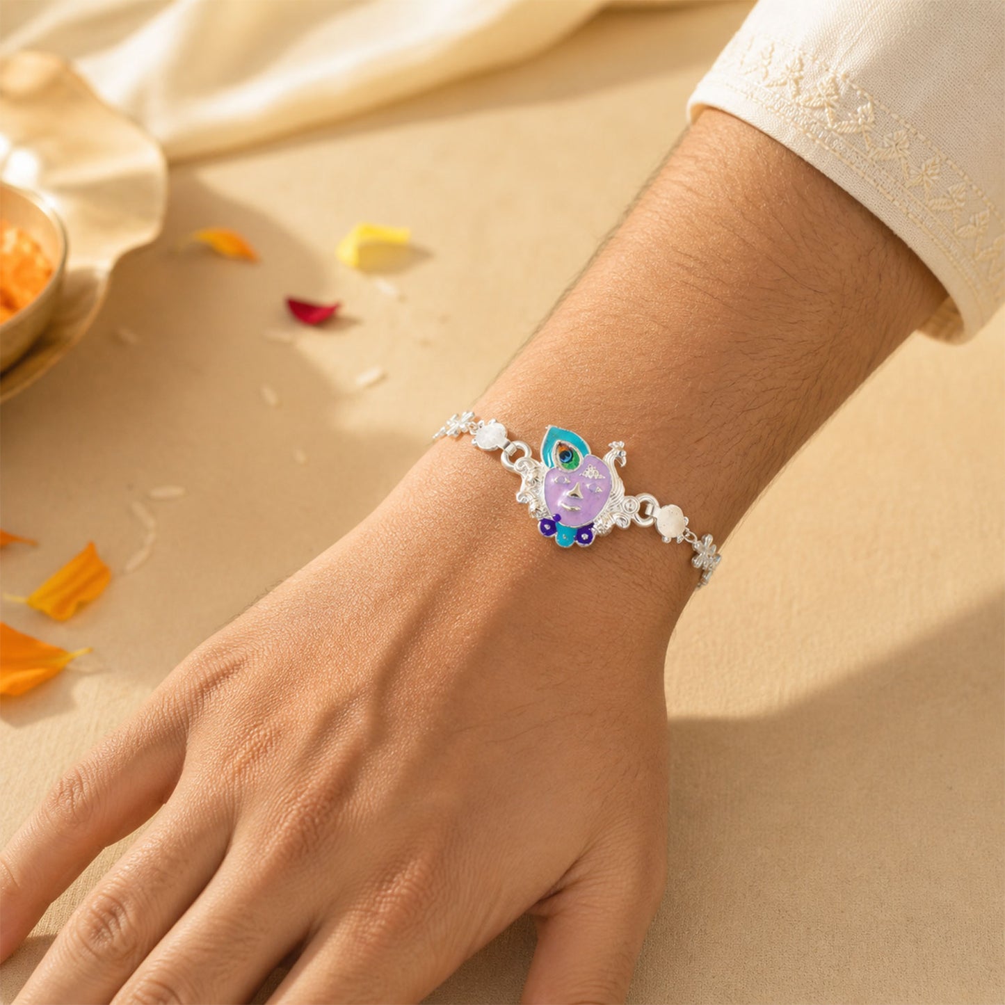 A person's wrist wearing the silver rakhi with a purple enamel Krishna face charm, surrounded by scattered petals.