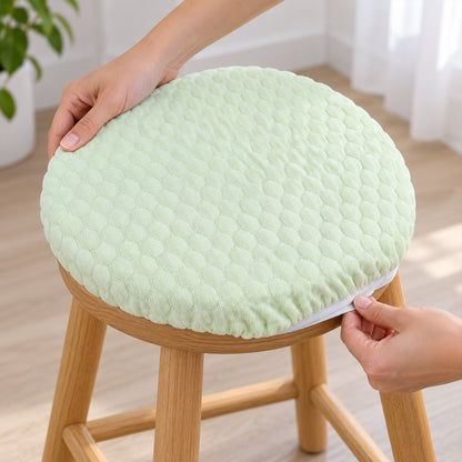 Hands adjusting the light green textured round cushion cover with an elastic edge onto a wooden stool.