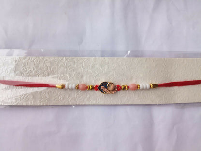 A red thread with a gold and blue peacock charm, flanked by pink, white, and gold beads, is displayed on a textured white