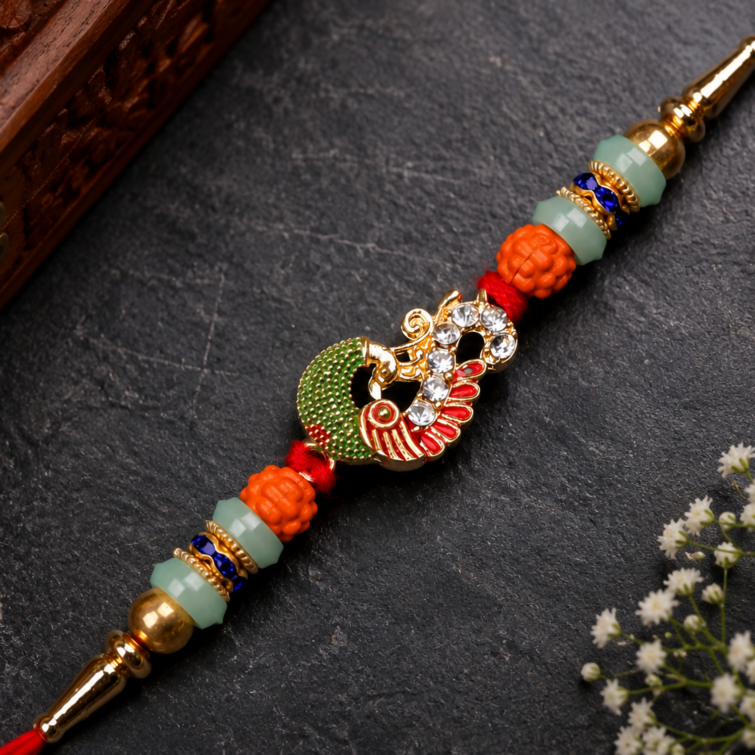 A red thread with a gold and green peacock charm, orange and mint green beads, and gold accents, rests on a dark gray