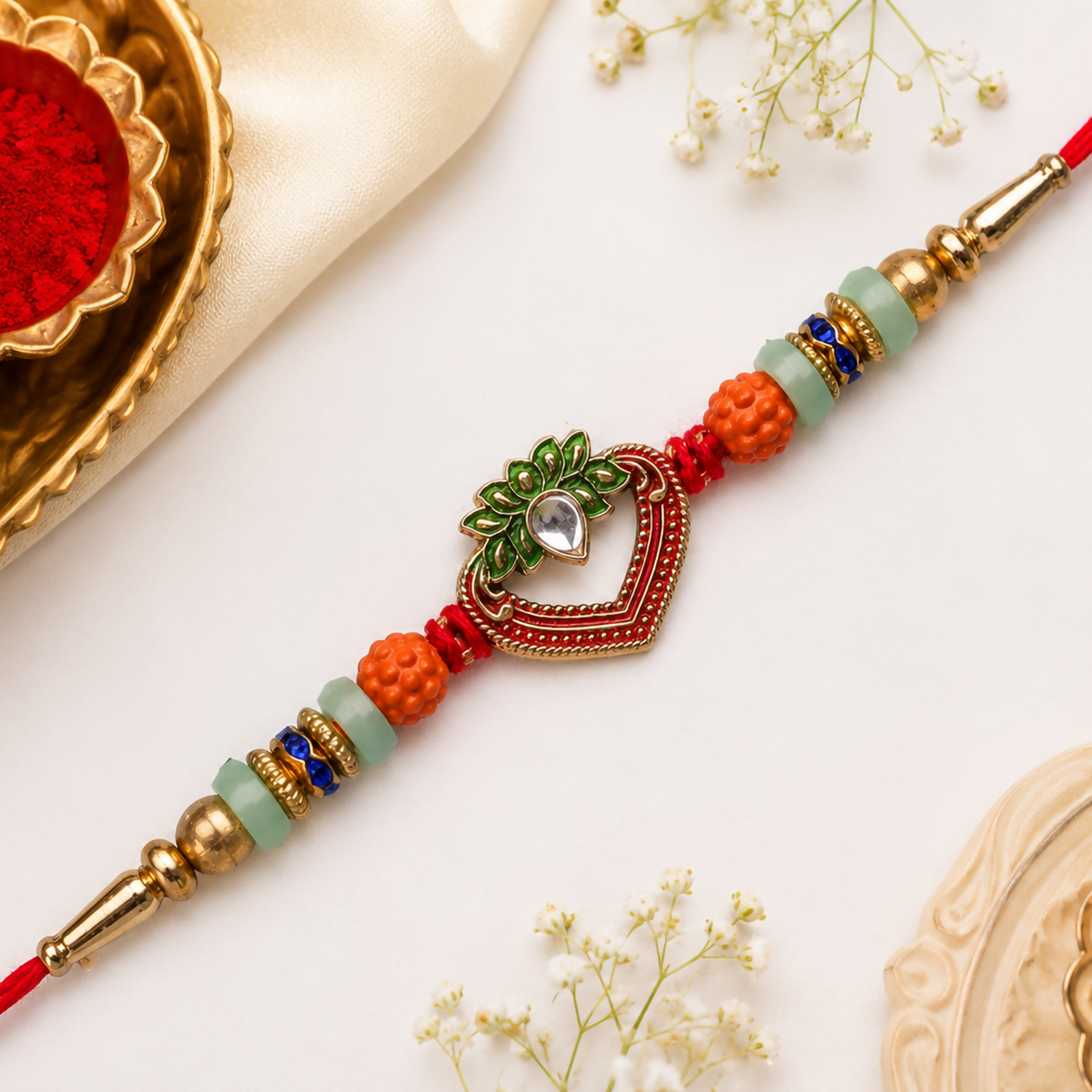 A red and gold bracelet with a heart-shaped charm, featuring green and orange beads, rests on a white surface with baby's