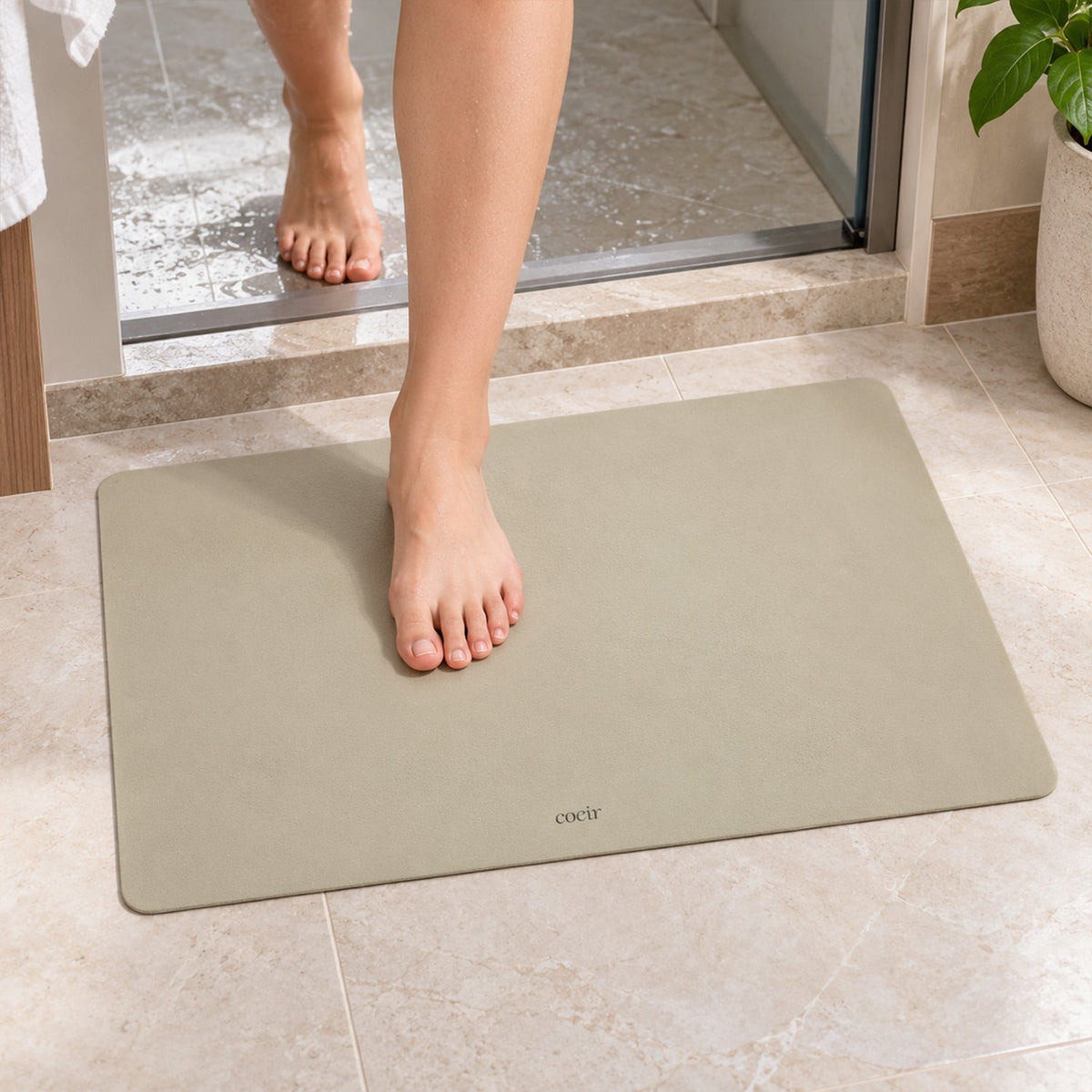 Bare feet step onto a light beige rectangular mat on tiled bathroom floor near a shower.