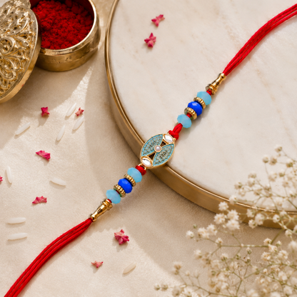 A red thread bracelet with blue and light blue beads and a golden oval charm with a light blue design, is placed on a light