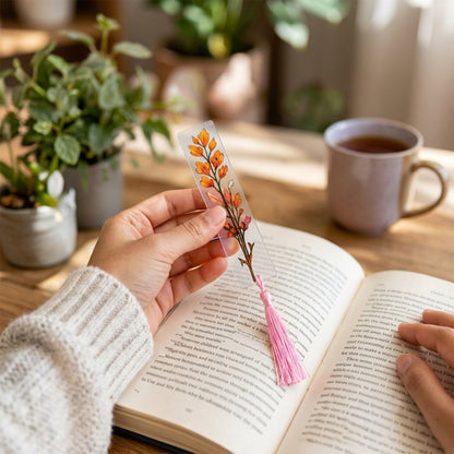 A hand holds a transparent bookmark with an orange leafy design and a pink tassel over an open book.