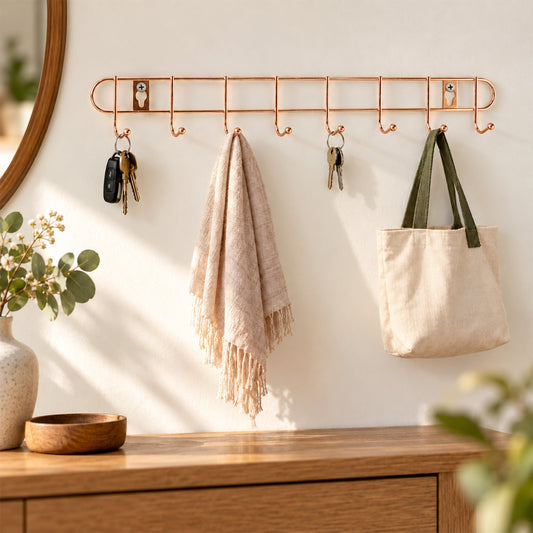 A rose gold metal wall hanger with eight hooks holds keys, a light pink towel, and a cream canvas bag.