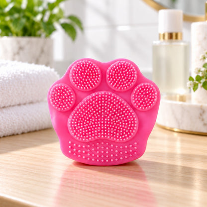 A vibrant pink paw-shaped facial scrubber with soft silicone bristles sits on a light wooden surface.