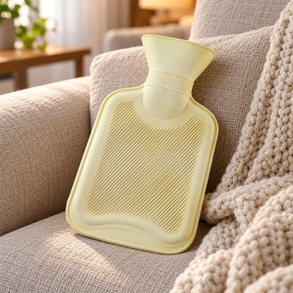 A light yellow hot water bottle with a ribbed surface rests on a beige upholstered armchair next to a knitted blanket.