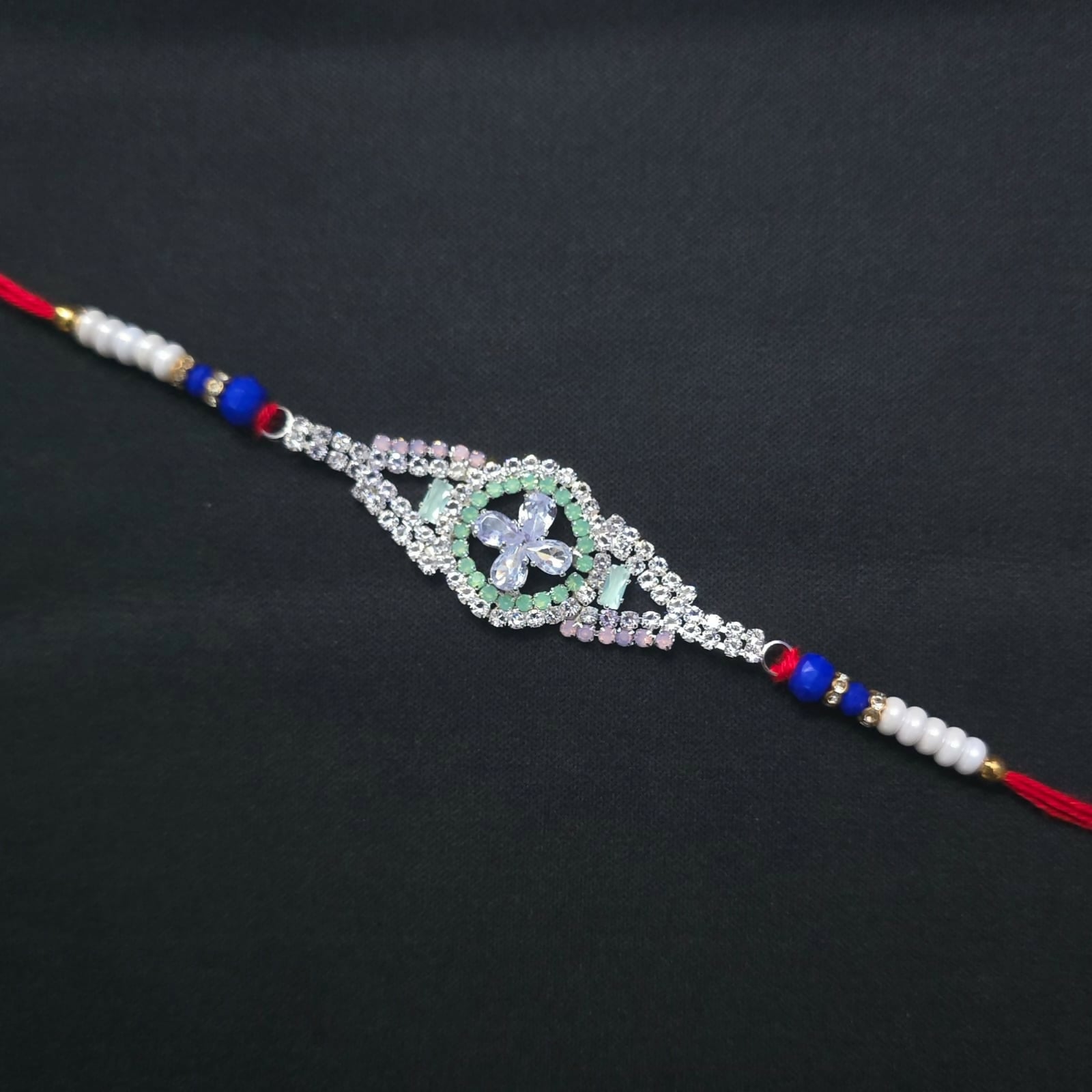 A single red thread bracelet with a prominent silver-toned, crystal-encrusted flower design and small white and blue beads,