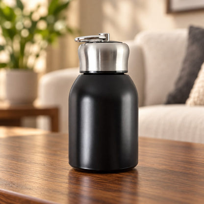 A matte black vacuum bottle with a shiny silver cap and carry ring rests on a polished wooden table.