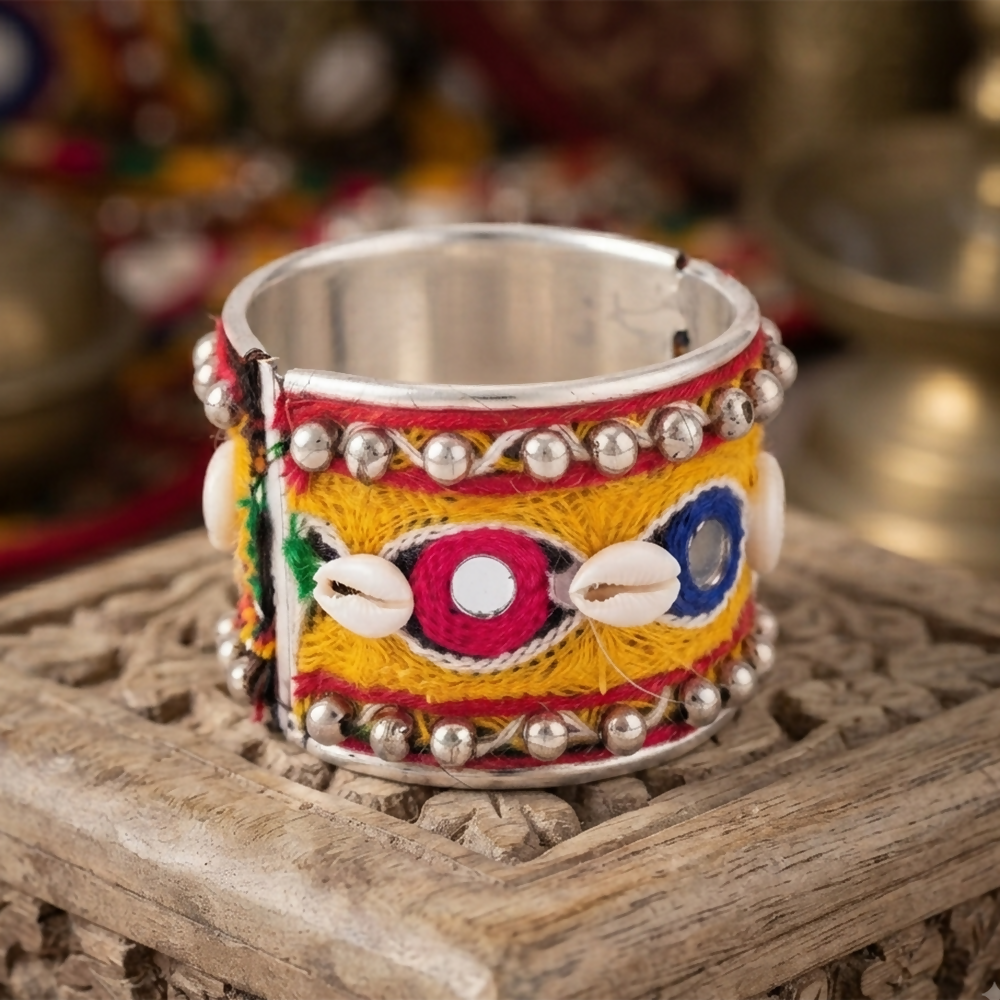 A vibrant metal cuff bracelet with yellow, red, and blue embroidery, small mirrors, and cowrie shells, resting on a carved